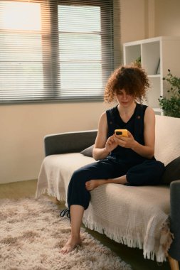 Beyaz kadın kanepede çıplak ayakla oturuyor ve sıcak bir sabah ışığında akıllı telefon kullanıyor..