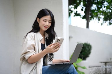 Açık kafede akıllı telefon ve laptop kullanan gülümseyen genç bir kadın..