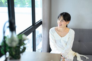 Asyalı kadın kanepede dinleniyor, neşeli bir ifadeyle pencereden dışarı bakıyor..