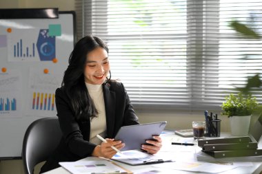 Gülümseyen iş kadını, modern bir ofiste dijital tablette finansal verileri analiz ediyor..