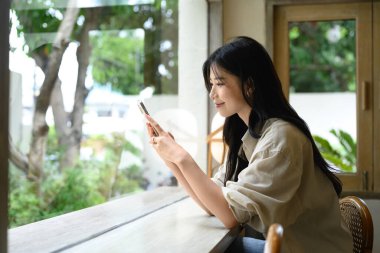 Gülümseyen genç bir kadın kafenin penceresinde oturmuş akıllı telefonunu kullanıyor..
