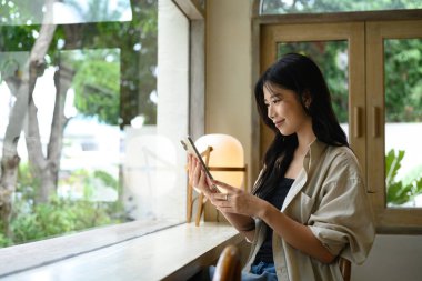 Gülümseyen Asyalı kadın şık bir kahve dükkanının penceresinde oturur ve akıllı telefon kullanır..