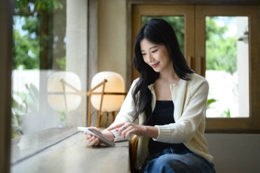 Gülümseyen Asyalı kadın şık bir kahve dükkanının penceresinde oturur ve akıllı telefon kullanır..