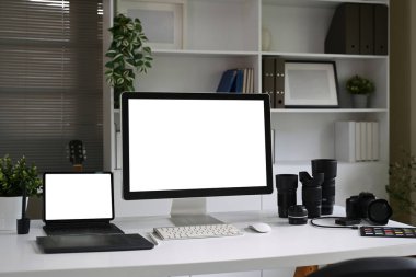 Yaratıcı stüdyodaki beyaz bir masada bilgisayar, tablet, kamera ve lenslerle fotoğrafçılık çalışma alanı.