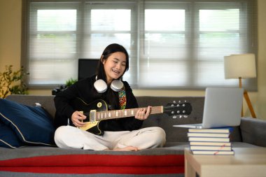 Evde elektro gitar çalan gülümseyen genç kız bilgisayardan sanal müzik dersi alıyor..