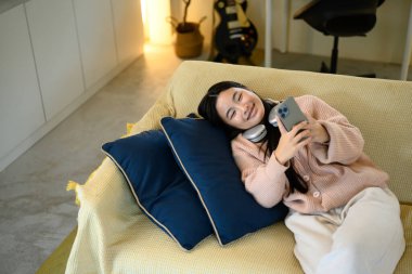 Neşeli genç kız kanepede uzanıyor, evde dinleniyor ve akıllı telefon kullanıyor..