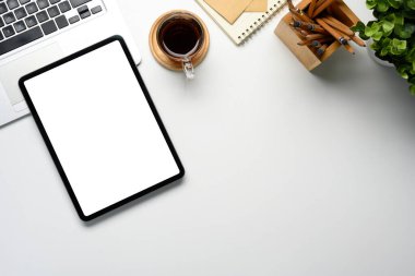 Boş ekranlı dijital tablet, kahve fincanı, dizüstü bilgisayar ve temiz beyaz bir masanın üzerindeki defter..