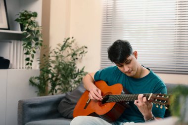 Genç beyaz bir adam kanepede oturup akustik gitar çalıyor..