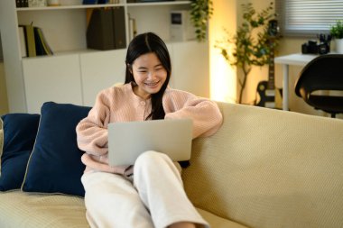 Gülümseyen genç bir kadın dizüstü bilgisayar kullanırken kanepede dinleniyor, online aktivitelerden zevk alıyor..