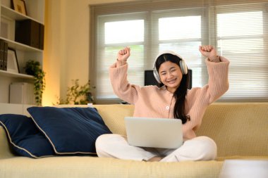 Kanepede dizüstü bilgisayarla oturan mutlu genç bir kadın çalışırken ya da internette çalışırken başarıyı kutluyor..