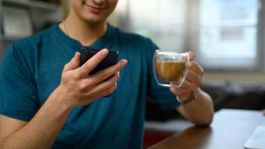 Elinde akıllı telefon ve bir bardak kahveyle oturma odasında oturan gülümseyen adama yaklaş..