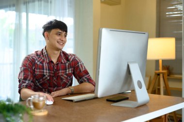 Modern bir ev ofisinde, masaüstü bilgisayarında çalışan gülümseyen bir adam..