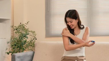 Gülümseyen genç bir kadın evdeki kanepede otururken koluna nemlendirici krem sürüyor..