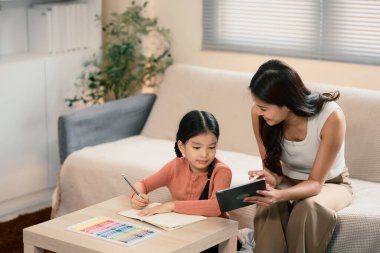 Anne kızına ödevlerinde yardım ediyor ve oturma odasında dijital tablet kullanıyor..