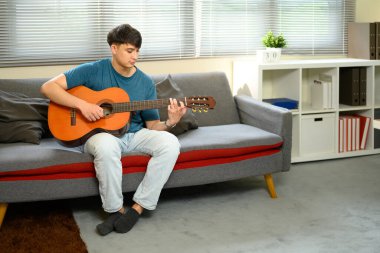 Genç bir adam parlak bir oturma odasındaki kanepede akustik gitar çalıyor..