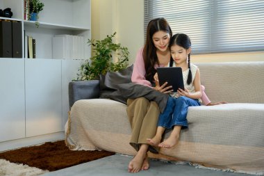 Anne ve kızı evde dijital tablet kullanarak bir kanepede oturuyorlar..