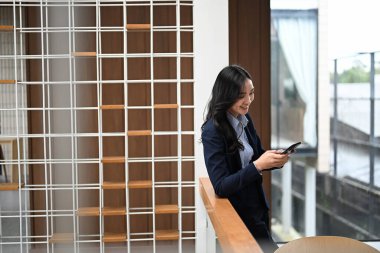 Gülümseyen iş kadını modern bir ofiste korkulukların yanında durup akıllı telefon kullanıyor..