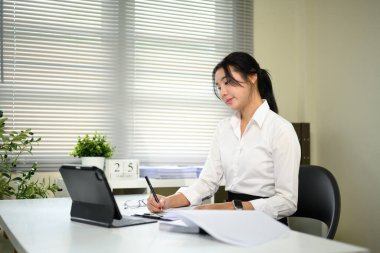 Modern bir ofiste dijital tablet üzerinde çalışırken not alan Asyalı iş kadını.