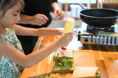 Küçük kız mutfakta sandviç yaparken ekmeğin üzerine mayonez sıkıyor..