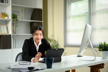 Asyalı iş kadını, ev ofisinin modern beyaz masasında dijital tablet kullanıyor..