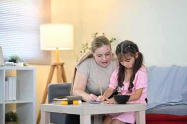 Bakıcı anne, kızına ev ödevlerinde yardım ediyor, birlikte oturma odasında oturuyor..