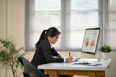 Masaüstü monitöründeki aylık bütçe çizelgelerini incelerken kağıda yazan kadın çalışan.