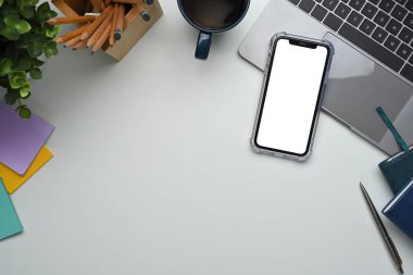 Boş ekran akıllı telefon, dizüstü bilgisayar, kahve fincanı ve ofis malzemeleri beyaz masada..