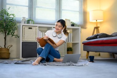 Genç bir kadın dizüstü bilgisayarla evde çalışırken elinde not defteriyle yerde oturuyor..