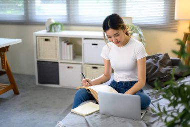 Evden çalışan genç bir kadın, kanepede dizüstü bilgisayar kullanırken deftere notlar yazıyor..