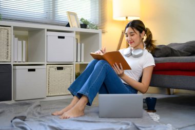 Gülümseyen genç bir kadın yerde kitap okuyor ve notlar alıyor..