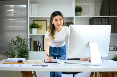 Genç girişimci masaüstü bilgisayarında çalışıyor ve modern ofiste notlar alıyor...