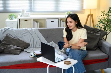 Küçük masada dizüstü bilgisayar kullanan Asyalı kadın, oturma odasında kahvenin tadını çıkarıyor..