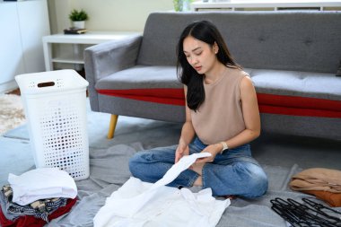 Genç bir kadın temiz çamaşırları evde katlıyor, yerde giysileri ve sepetle oturuyor..