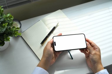 Masanın üzerinde boş beyaz ekran maketi ve defter ve kalemle akıllı telefon tutan eller.