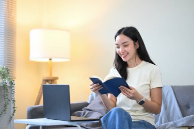 Gülümseyen genç kadın kanepede oturuyor, kitap okuyor ve dizüstü bilgisayarla oturma odasında ders çalışıyor..