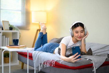 Kanepede kulaklıkla uzanan ve müzik ya da video yayını için akıllı telefon kullanan genç bir kadın..