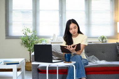 Sıradan bir kadın evde dizüstü bilgisayarla çalışırken kitap okuyor ve not alıyor..