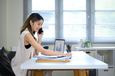 Sırıtkan iş kadını, bilgisayar ve belgelerle çalışırken telefonda konuşuyor..