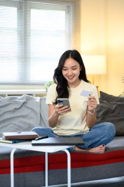 Kanepede oturan mutlu kadın elinde cep telefonu ve kredi kartıyla internetten alışveriş yapıyor..