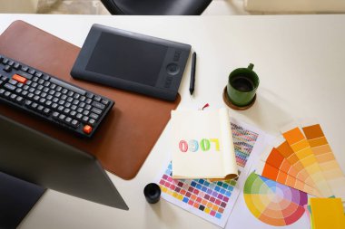 Top view of graphic designer workspace with logo sketch, color palette, and drawing tablet on white desk.