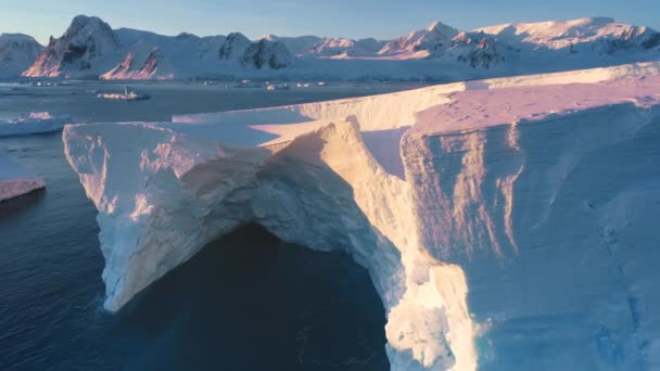 巨大な氷河のアーチが南極に墜落した ピンクの日没灯の巨大な氷山の壁 北極の凍った海の風景 雪が背景の山々を覆いました トラベル エアリアルビュードローンフライトズーム — ストック動画