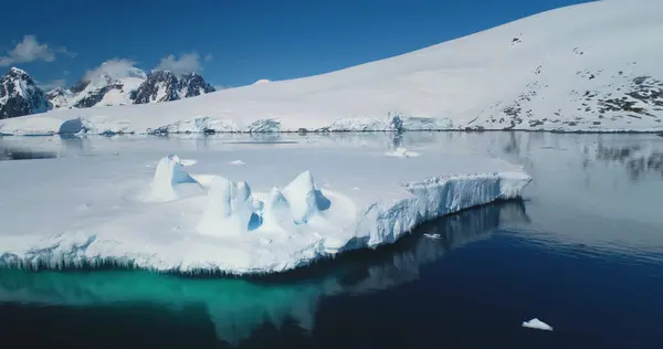 Arctic winter landscape at global warming problem. Glacier icicles melting in blue ocean water, snow covered hill against blue sky in sunny day. Antarctica polar summer. Drone flight close up panorama