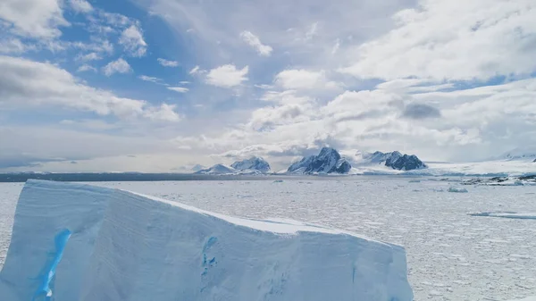 Aerial Flight Over Antarctica Iceberg, Ocean. White Landscape. Amazing Drone Shot Of Ice Frozen Ocean. Cloudy Sky And Sunlit Snow Covered Mountains Background. Winter South Pole Scenery. 4k Footage.