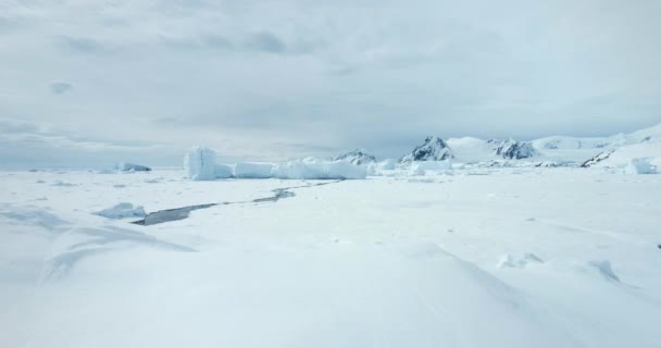 南極の冬の風景に雪の丘の上を飛んでください 溶けたひび割れた川で凍った極海 氷河と氷山が氷の海岸湾に閉じ込められた バックグラウンドの山脈 北極圏の旅と探検 — ストック動画