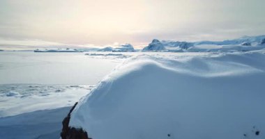 Gün batımında donmuş Antarktika karlarının üzerinden uç. Güney Kutbu 'nun el değmemiş vahşi doğası. Çöl beyazı manzarası, kar ve buz gibi hava aracı görüntüleri. Arka planda dağlar. Doğanın kutup koruması