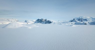 Kuzey Kutbu 'ndaki kış manzarası karların üzerinde uçmak, arka planda yükselen dağlar. Güneşli bir günde mavi gökyüzü altında donmuş Antarktika manzarası. Kutup donmuş okyanusu. Düşük açılı hava aracı atışı
