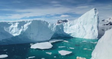 Eriyen buzdağları turkuaz su okyanusu koyunda yüzer. Arka planda karlı dağlar var. Antarktika 'da mavi gökyüzünün altındaki dev buzullar. Küresel ısınma, eriyen buz, iklim değişikliği. İHA atışı. Yakın çekim.