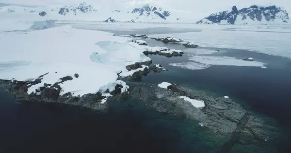 Antarktika 'daki kayalık kıyı şeridi karla kaplıydı. Soğuk sularda kayalar, arka planda dağlar olan donmuş kutup okyanus körfezi. Antarktika seyahati ve keşfi. Hava aracı görüntüsü