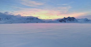 Winter sunset snowy landscape with towering mountains range in background. Fly over untouched wilderness Antarctica snow covered arctic nature. Polar frozen land under bright colorful sky. Drone shot
