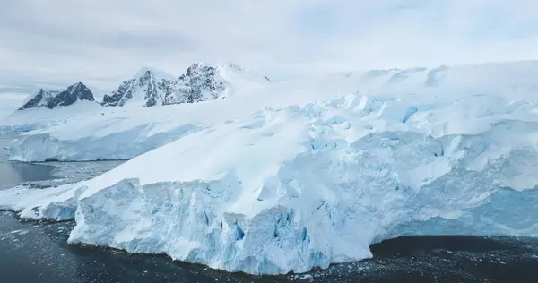 stock image Blue snowy glacier towering Antarctica coastline. Snow covered giant iceberg ice wall rises above cold, dark ocean water. Antarctic winter polar landscape panorama. Aerial cinematic ecology scene